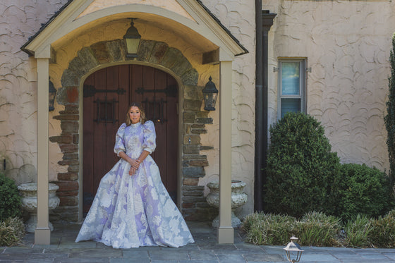 Woman wearing a modest lilac purple ballgown featuring a white and silver floral overaly with a high neckline and three quarter sleeve puff sleeves.