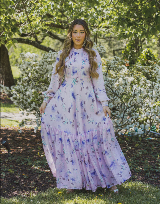 A woman wearing a modest light pink gown with lilac florals and a large ruffle hem.