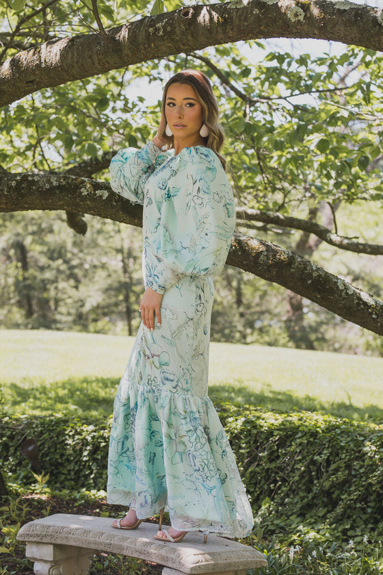 Woman wearing modest mint green gown with a floral print. The gown has full, voluminous sleeves and a ruffled bottom hem that adds a playful touch. 