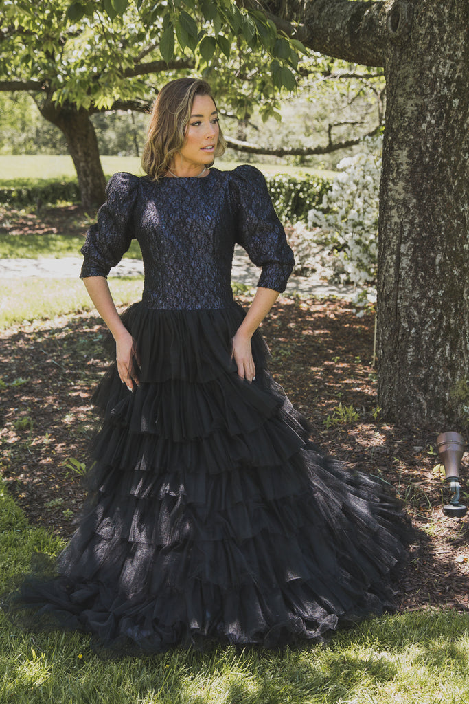 Woman wearing modest black tiered ball gown. The gown features three-quarter sleeves with puff sleeve detail. 