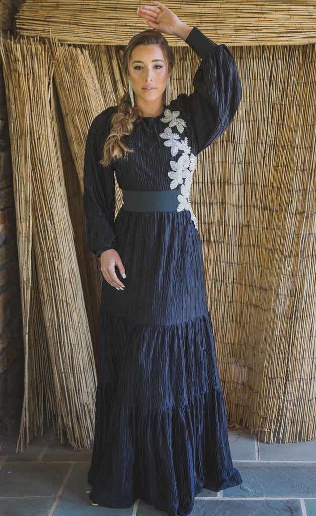 Woman wearing a black modest boho style gown with long sleeves and white beaded flowers cascading down one side.