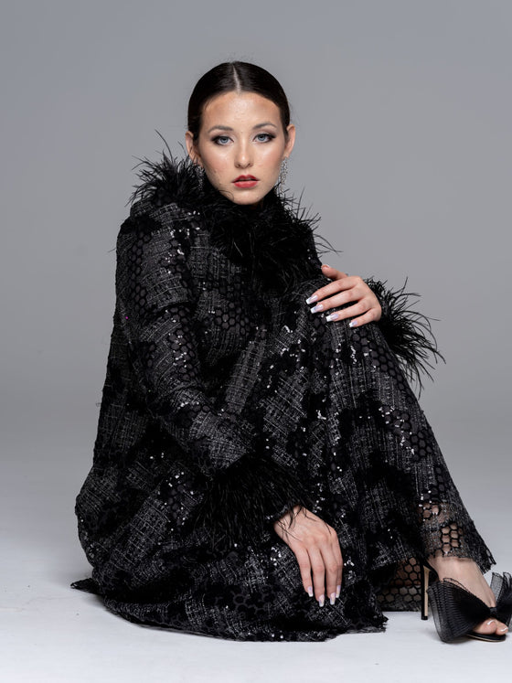 A woman sitting and wearing a black fitted modest gown with a high neckline and long sleeves. The gown has sequin detailing and a black feather collar and cuffs. 