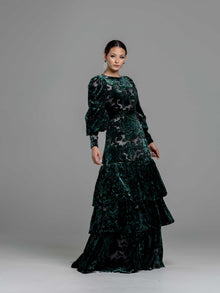  A woman wearing a modest dark green floral velvet burnout gown with puff sleeves. The gown has a fitted bodice, the bottom of the gown has 3 tiers of ruffles. 