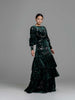 A woman wearing a modest dark green floral velvet burnout gown with puff sleeves. The gown has a fitted bodice, the bottom of the gown has 3 tiers of ruffles. 