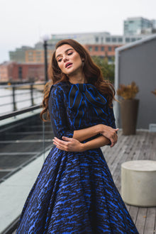  Woman wearing modest cobalt blue and black ball gown with cropped bolero. The three-quarter sleeve bolero has a large bow detail in front and mini covered buttons cascading down the back.