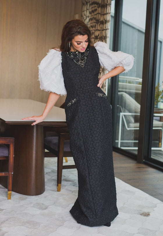 A woman wearing a modest straight black gown with contrasting three-quarter sleeve white balloon sleeves and pockets. The gown is adorned with exotic beading in a triangle by the neck and a line of beading on the pockets.