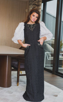  A woman wearing a modest straight black gown with contrasting three-quarter sleeve white balloon sleeves and pockets. The gown is adorned with exotic beading in a triangle by the neck and a line of beading on the pockets.