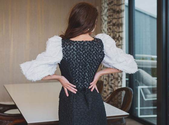 A woman wearing a modest straight black gown with contrasting three-quarter sleeve white balloon sleeves and pockets. The gown is adorned with exotic beading in a triangle by the neck and a line of beading on the pockets.