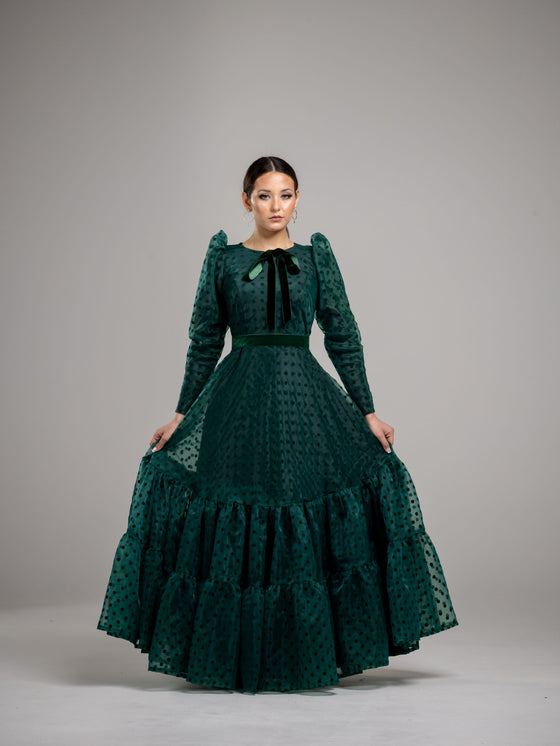 Woman wearing an elegant modest hunter green gown with velvet polka dots, a bottom tier and a green velvet bow at the neck. 