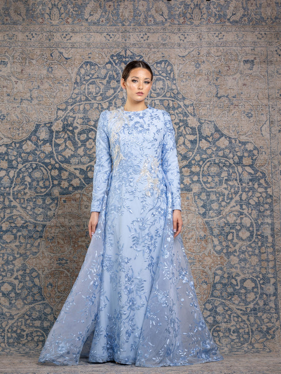 Woman wearing a modest powder blue floral long sleeve gown with a flowing skirt cape for added elegance.