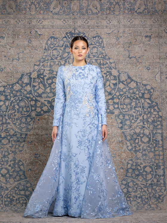 Woman wearing a modest powder blue floral long sleeve gown with a flowing skirt cape for added elegance.