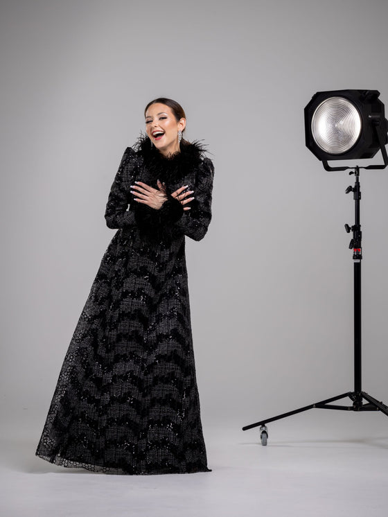 A smiling woman wearing a black fitted modest gown with a high neckline and long sleeves. The gown has sequin detailing and a black feather collar and cuffs. 