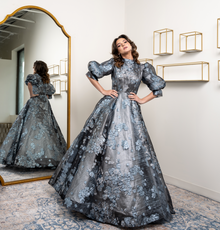  A woman wearing a modest metallic silver gown with a silver and blue burnout floral pattern and three quarter sleeve balloon sleeves.