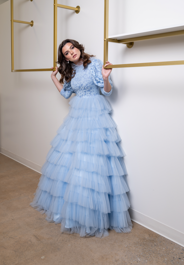 A woman wearing a modest powder blue ball gown. The skirt features layers of accordion tulle. The three-quarter  voluminous sleeves are cut in a draped fabric allowing for maximum movement. The top is floral printed and has a high collar.