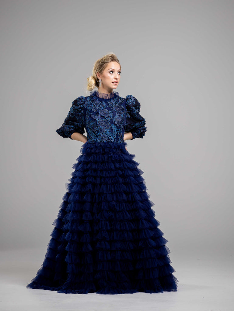 A woman wearing a navy blue modest ball gown with a floral top and high neckline. The gown has a fitted bodice and a flowing skirt with tiers of lace.