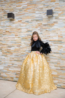  A girl wearing a modest black and gold two-piece ball gown with rich black feathers cascading down one sleeve and an imported gold crinkle foiled fabric ball gown skirt. 