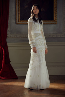  Woman wearing a modest ivory Needle & Thread gown.  The gown is crafted from vintage tulle and lace and adorned with intricate beaded embellishments.  It has a fitted bodice, fluted sleeves and a floor sweeping hem.