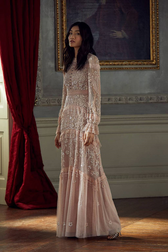 Women wearing modest blush pink vintage tulle and lace Needle & Thread gown. The gown is adorned with beaded embellishments and outlined in ruffles. The gown features a fitted bodice, fluted sleeves, and a floor sweeping hem.