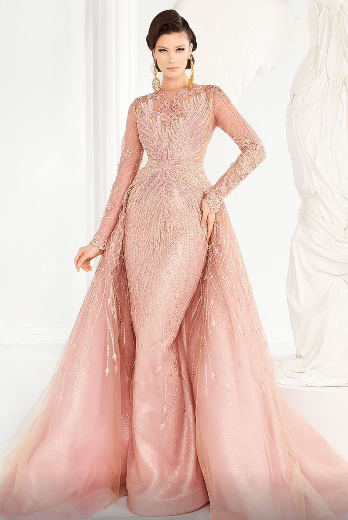 A woman wearing a pink, modest gown crusted with tonal beadwork of an intricate design. The embellished long sleeves add a touch of glamour, and the mermaid skirt gives the gown a sleek and sophisticated silhouette. The full-length gown is finished with a soft, open overskirt that flares from the waist to a court train, adding a touch of drama to the overall look.