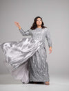 A woman wearing a silver modest straight silhouette gown adorned with sparkling sequins and a skirt cape. The gown has a high neckline and long sleees. 