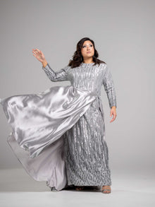  A woman wearing a silver modest straight silhouette gown adorned with sparkling sequins and a skirt cape. The gown has a high neckline and long sleees. 